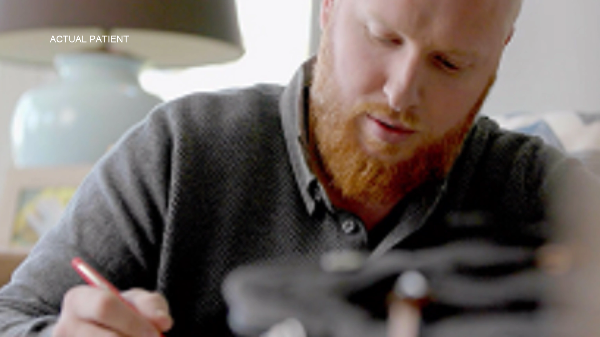A man wearing a gray T-shit with a beard is making notes using a pen with a bed lamp in the background.