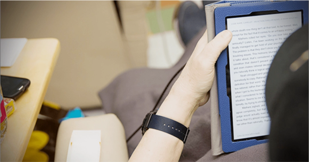 A person wearing a black watch holding a tablet displaying text, with various items in the background.