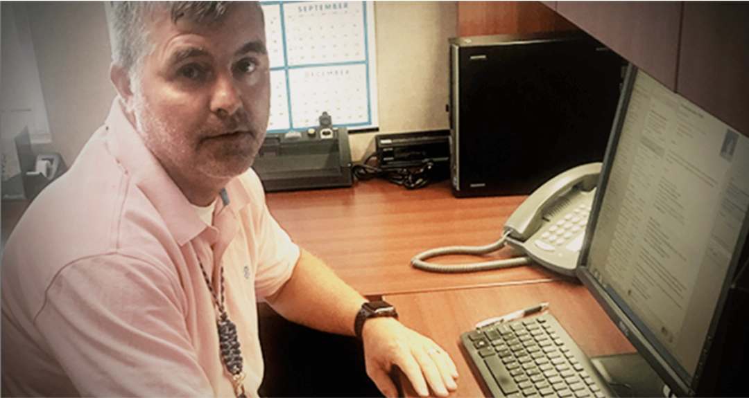 A man with short gray hair wearing a pink shirt is seated in an office environment.