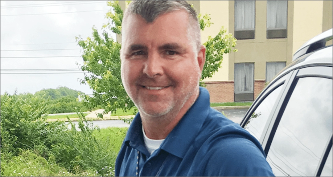 A man with short gray hair and a blue shirt stands near a car with a building and greenery in the background.