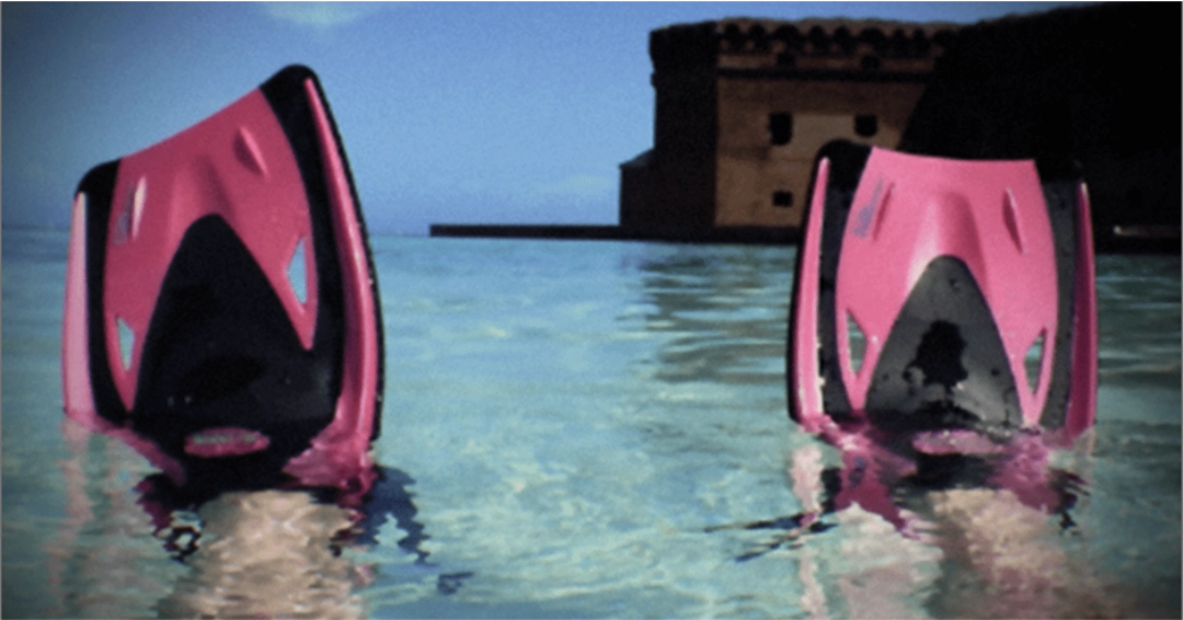 Two pink and black swim fins partially submerged in clear blue water, with a fort visible in the background.