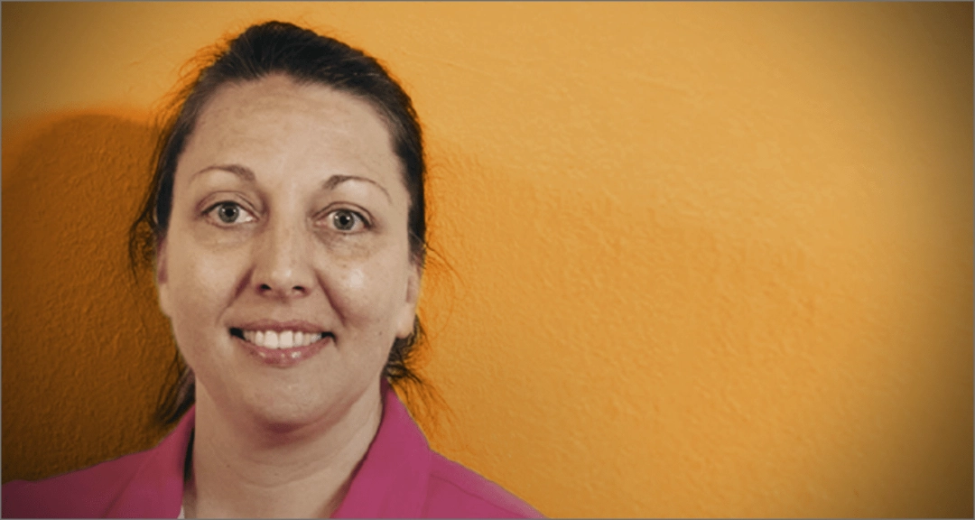 A woman wearing a pink shirt stands in front of an orange background.