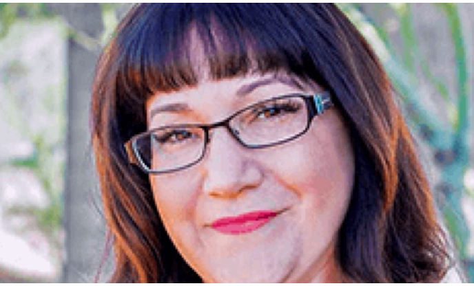 A woman is wearing glasses, with greenery in the background.