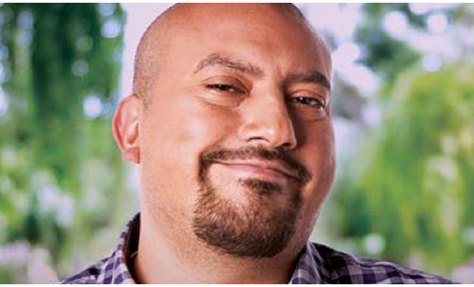 A man with a beard wearing a checked shirt, with greenery in the background.