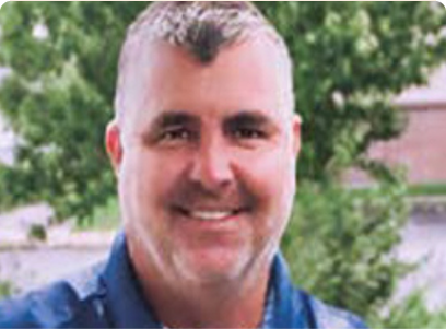 A man with short gray hair wearing a blue shirt, with greenery in the background.