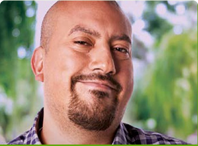 A man with a beard wearing a checked shirt, with greenery in the background.