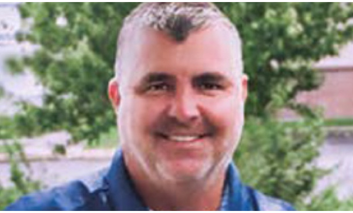 A man with short gray hair wearing a blue shirt, with greenery in the background.